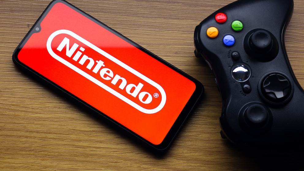 Ein Smartphone mit rotem Nintendo-Logo liegt auf einem Holztisch. Daneben befindet sich ein schwarzer Spielecontroller mit farbigen Tasten.