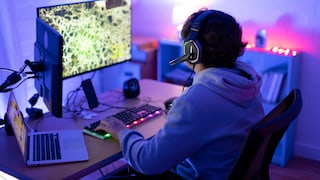 Auf dem Bild sitzt eine Person mit Headset an einem Schreibtisch vor einem großen Monitor und spielt offenbar am PC. Die Szene ist vor allem in blauem und violettem Licht gehalten. Vor der Person stehen eine beleuchtete Tastatur und weiteres Computerzubehör. Links befindet sich zusätzlich ein aufgeklappter Laptop. Im Hintergrund sind Möbel und weitere Lichtquellen zu erkennen.