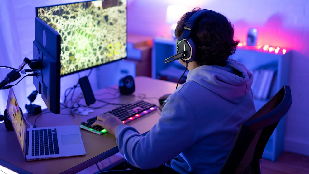 Auf dem Bild sitzt eine Person mit Headset an einem Schreibtisch vor einem großen Monitor und spielt offenbar am PC. Die Szene ist vor allem in blauem und violettem Licht gehalten. Vor der Person stehen eine beleuchtete Tastatur und weiteres Computerzubehör. Links befindet sich zusätzlich ein aufgeklappter Laptop. Im Hintergrund sind Möbel und weitere Lichtquellen zu erkennen.