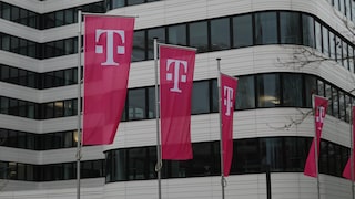 Auf dem Bild stehen mehrere magentafarbene Telekom-Flaggen mit dem weißen „T“-Logo vor einem modernen Bürogebäude. Weitere Flaggen sind am rechten Bildrand zu sehen, im Hintergrund erstreckt sich die helle Fassade mit großen dunklen Fensterflächen.