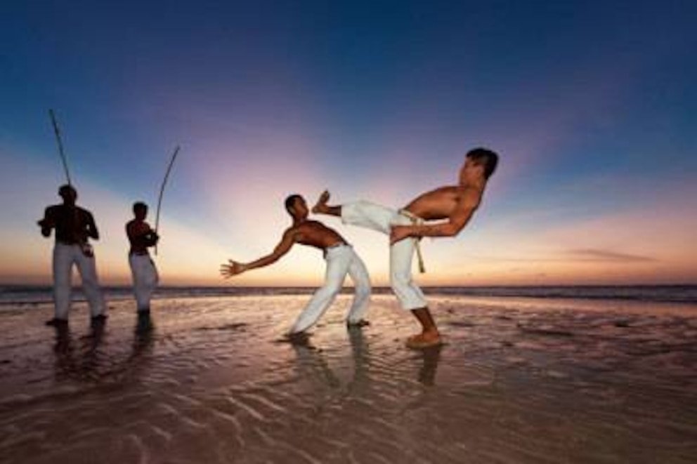 Capoeira-Kämpfer am Strand: Das Instrument, das die Männer im Hintergrund spielen, heißt Berimbau und gibt den Rhythmus und damit die Geschwindigkeit des Kampfes, Spiel genannt, vor