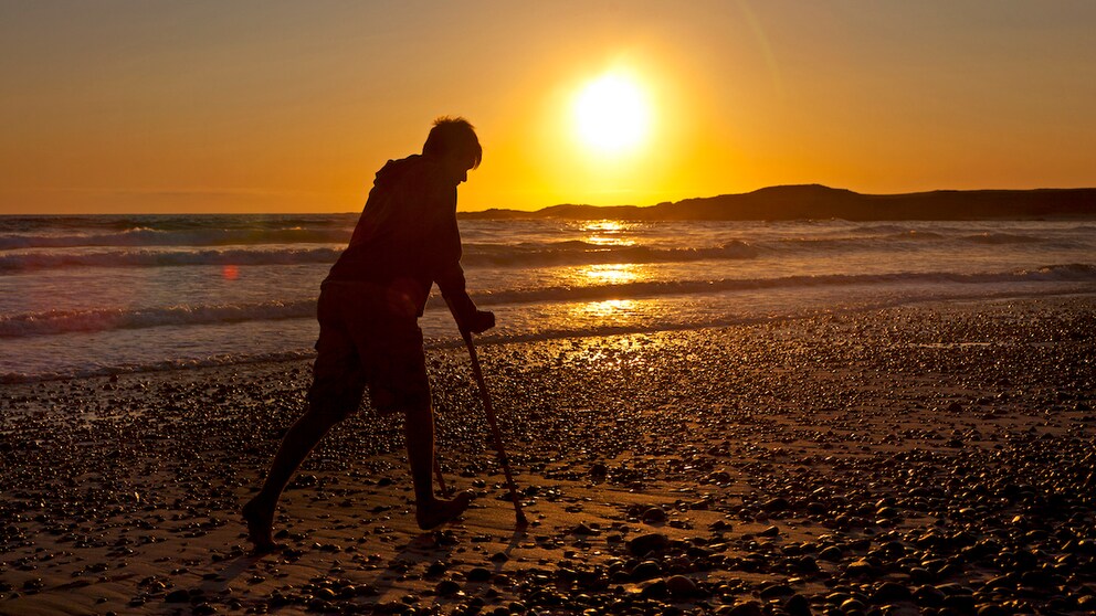 Auslandskrankenversicherung: Junger Mann geht mit krücken am Strand im Sonnenuntergang