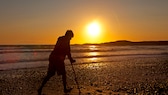 Auslandskrankenversicherung: Junger Mann geht mit krücken am Strand im Sonnenuntergang