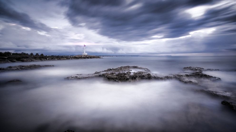 Mitten im Meer taucht plötzlich eine kleine, dicht bewaldete Insel auf. Ihre Küste fällt in schroffen Felswänden steil ab, der Himmel über ihr ist von dunklen Wolken verhangen.