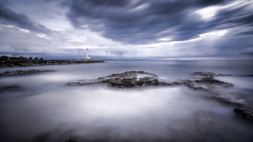 Mitten im Meer taucht plötzlich eine kleine, dicht bewaldete Insel auf. Ihre Küste fällt in schroffen Felswänden steil ab, der Himmel über ihr ist von dunklen Wolken verhangen.